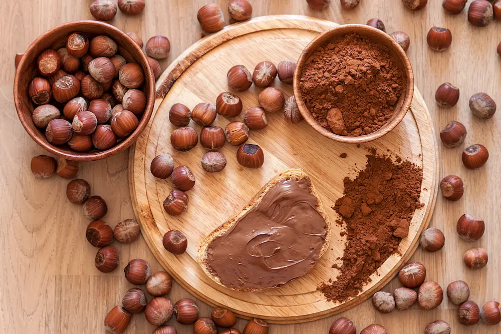 Bread with our hazelnut spread Cutting board with hazelnuts, cocoa, and bread spread with hazelnut cream